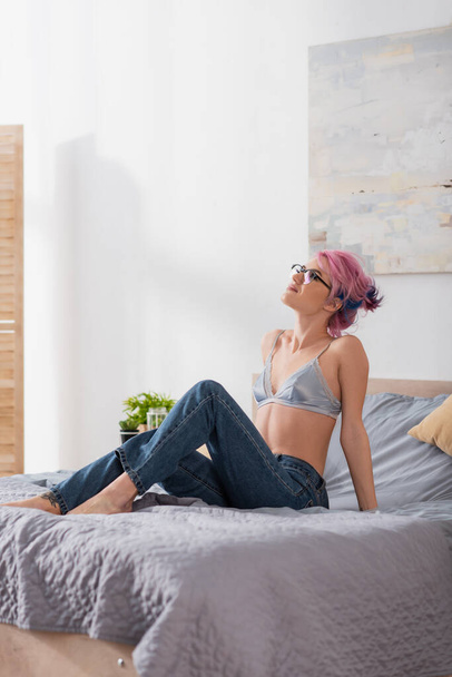 mujer joven soñadora con el pelo teñido sentado en sujetador de seda y jeans en la cama - Foto, imagen