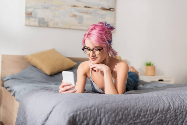 cheerful young woman with dyed hair lying on bed while messaging on smartphone - Photo, image