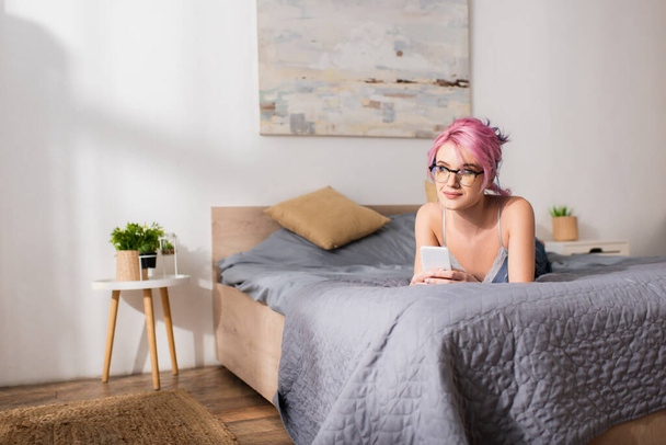 álmodozó fiatal nő festett hajjal fekszik az ágyon, miközben gazdaság smartphone - Fotó, kép