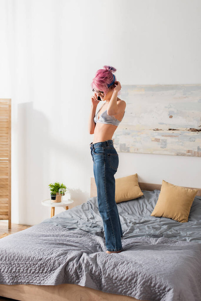 smiling young woman with dyed hair standing on bed while talking on smartphone - Foto, Imagen