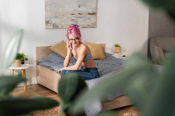 sonriente mujer joven con el pelo teñido sentado en la cama mientras habla en el teléfono móvil - Foto, Imagen