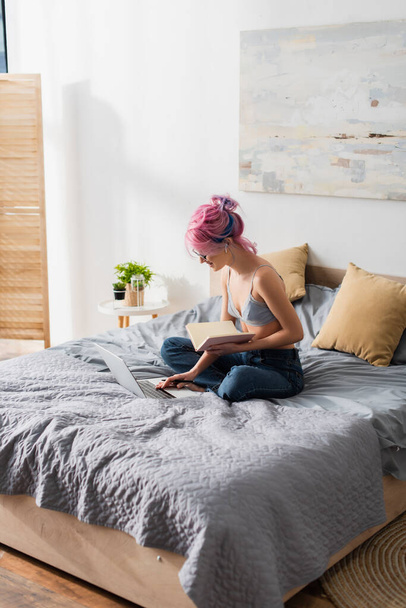cheerful woman with dyed hair holding notebook while using laptop and studying online at home - Foto, Imagem
