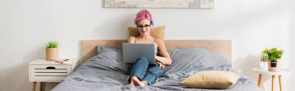 usmívající se žena s obarvenými vlasy ležící v podprsence a džínách při sledování filmu na notebooku - Fotografie, Obrázek