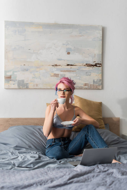 young woman with pink hair and earphone holding cup of coffee near laptop on bed - Фото, изображение