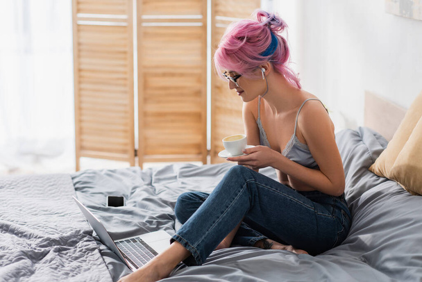 smiling young woman in earphone holding cup of coffee near gadgets on bed - Foto, afbeelding