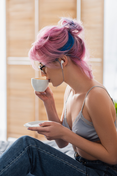 πλαϊνή όψη νεαρής γυναίκας με βαμμένα μαλλιά και ακουστικό πίνοντας καφέ - Φωτογραφία, εικόνα