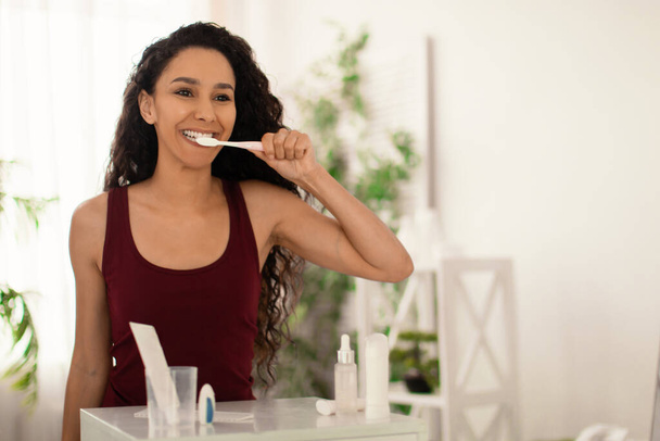 Pretty young woman brushing teeth with toothpaste near mirror at home, copy space - Photo, Image