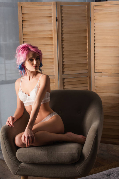 sexy jovem mulher com cabelo tingido sentado em poltrona perto de tela dobrável - Foto, Imagem