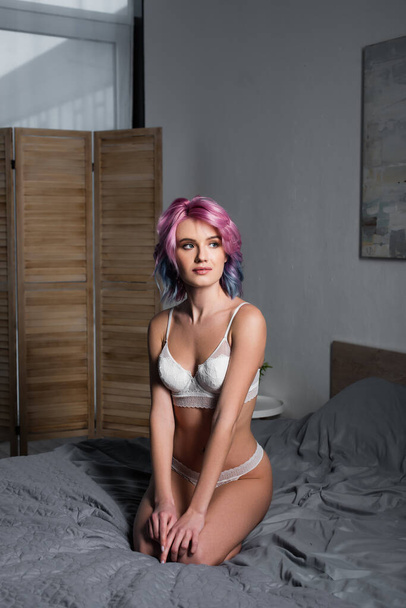 mujer joven en ropa interior blanca mirando hacia otro lado mientras está sentada en la cama - Foto, imagen