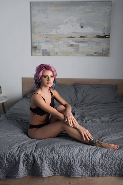 wytatuowana i seksowna kobieta z farbowanymi włosami w ciemnej koronkowej bieliźnie siedząca na łóżku - Zdjęcie, obraz