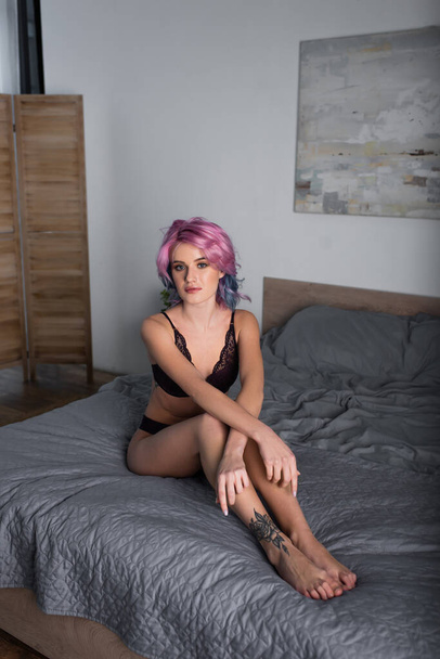 tetovált és szexi nő festett haj sötét fehérnemű ül az ágyon - Fotó, kép