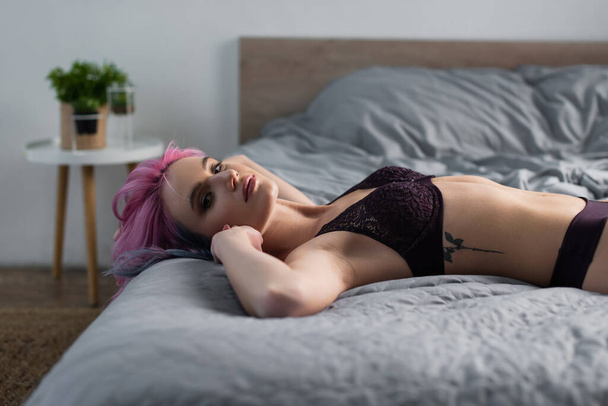 wytatuowana i seksowna kobieta w ciemnej bieliźnie patrząca w kamerę leżącą na łóżku - Zdjęcie, obraz