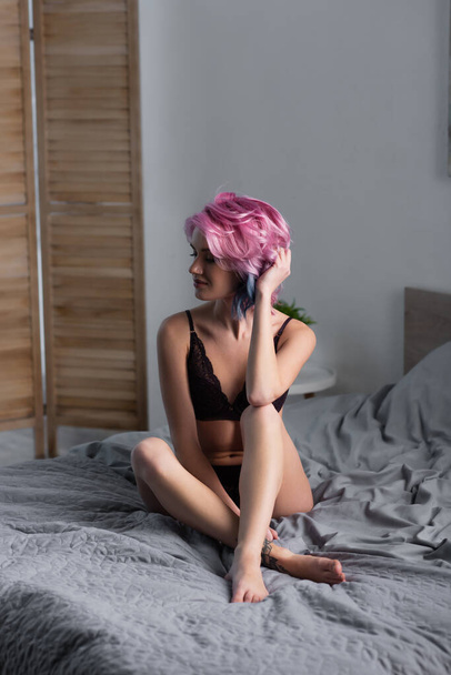 Genç, çıplak ayaklı, siyah iç çamaşırlı kadın yatak odasında oturuyor. - Fotoğraf, Görsel