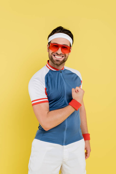 Pozitivní sportovec ve slunečních brýlích ukazuje svaly izolované na žluté  - Fotografie, Obrázek
