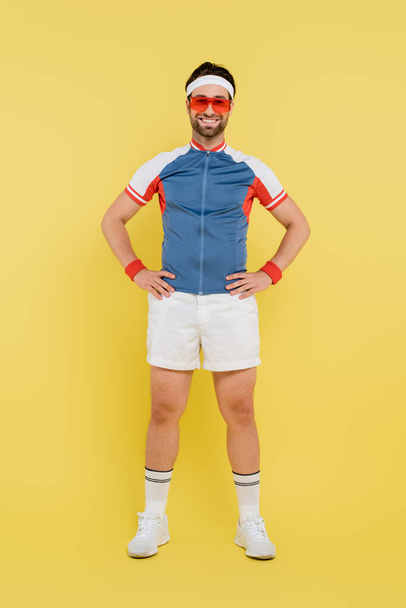 Full length of smiling sportsman in sunglasses holding hands on hips on yellow background  - Valokuva, kuva