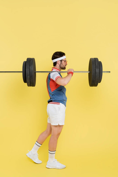 Side view of sportsman in sunglasses holding barbell on shoulder on yellow background  - Foto, Imagem