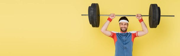 Young sportsman lifting barbell and looking at camera isolated on yellow, banner  - Valokuva, kuva