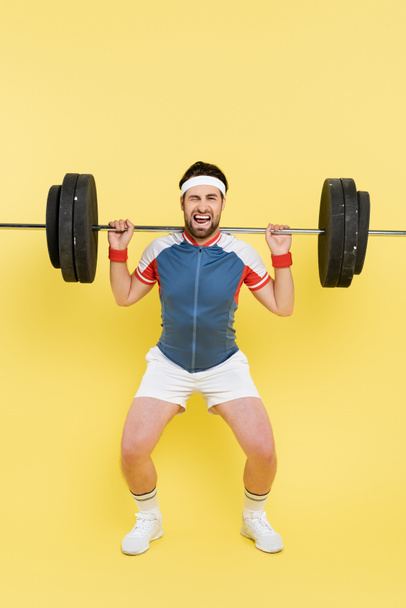 Young sportsman grimacing while lifting barbell on yellow background  - Foto, Imagem
