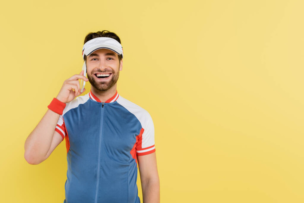 Hombre positivo en ropa deportiva y visera hablando en celular aislado en amarillo  - Foto, imagen
