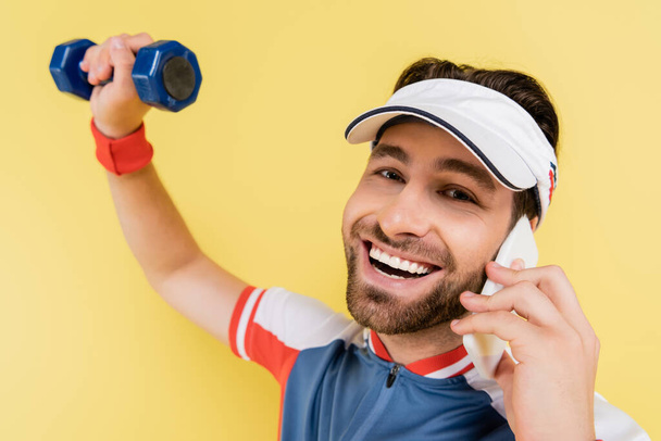 Uśmiechnięty sportowiec rozmawiający na smartfonie i patrzący w kamerę podczas treningu z hantlami odizolowanymi na żółto  - Zdjęcie, obraz