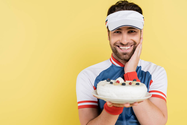 Positive sportsman in visor holding blurred cake isolated on yellow  - 写真・画像