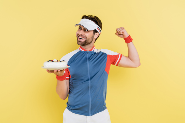 Young sportsman showing muscles and holding cake isolated on yellow  - Foto, immagini