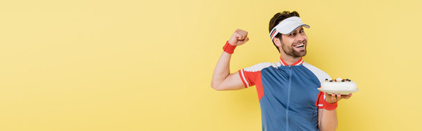 Cheerful sportsman holding cake and showing muscles isolated on yellow, banner  - 写真・画像