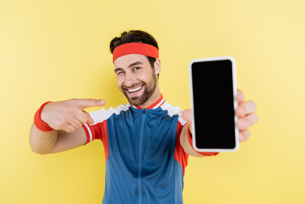 Smiling sportsman winking and pointing at blurred smartphone with blank screen isolated on yellow  - Fotó, kép
