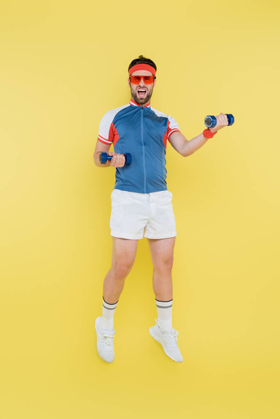 Sportsman in occhiali da sole saltando e tenendo i manubri isolati sul giallo  - Foto, immagini