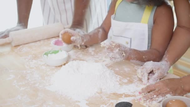 Close-up handen van Afro-Amerikaanse familie met dochter ei toevoegen aan bloem en dorsen voor het koken met vader en moeder samen in de keuken thuis, ouder en klein kind bereiden van voedsel met plezier. - Video
