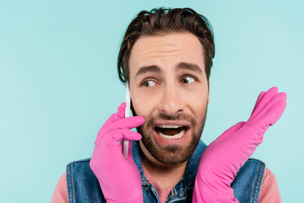 Young man in rubber gloves talking on mobile phone isolated on blue  - Foto, imagen