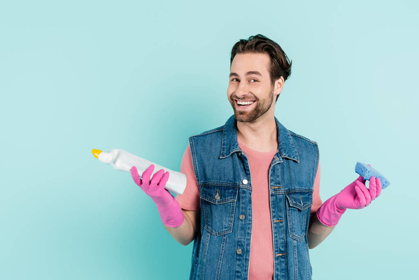 Positive young man in rubber gloves holding sponge and detergent isolated on blue  - Foto, imagen