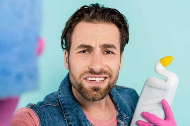 Młody człowiek w gumowych rękawiczkach trzyma gąbkę i detergent odizolowany na niebiesko  - Zdjęcie, obraz