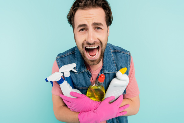 Young unmarried man in rubber gloves holding detergents and screaming isolated on blue  - Φωτογραφία, εικόνα