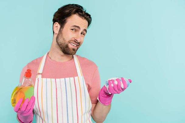 Sad man in apron and rubber gloves holding dishwashing liquid and sponge isolated on blue  - Foto, Imagen