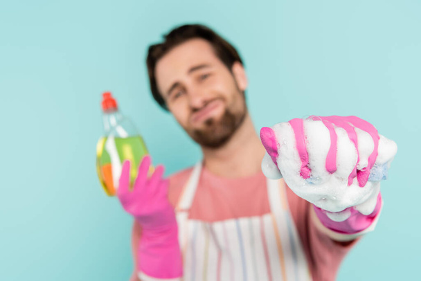 Hombre borroso con guantes de goma que sostienen detergente y esponja con espuma aislada en azul  - Foto, imagen