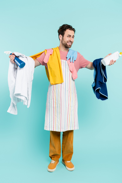 Piena lunghezza di uomo sorridente in grembiule che tiene detergenti e vestiti su sfondo blu  - Foto, immagini