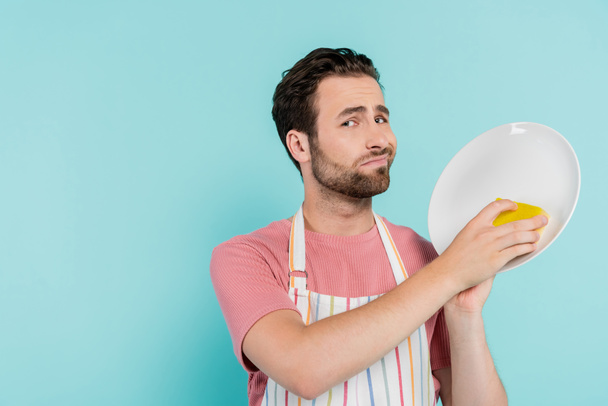 Nespokojený muž v zástěře čistící talíř s houbou izolovanou na modré  - Fotografie, Obrázek