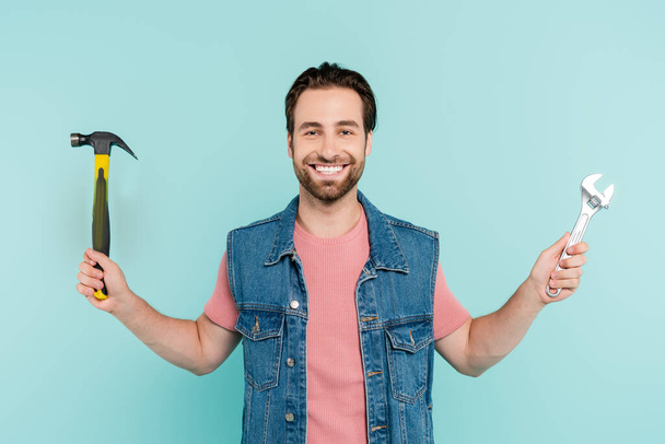 Cheerful man in denim vest holding hammer and wrench isolated on blue  - Fotoğraf, Görsel