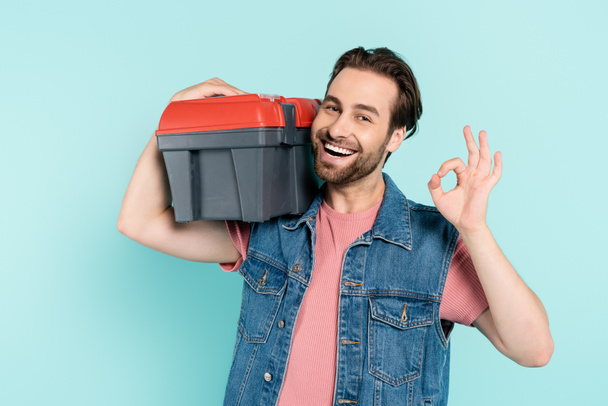 Cheerful man in denim vest holding toolbox and showing ok gesture isolated on blue  - Foto, Imagen