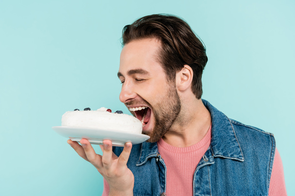 Młody mężczyzna w dżinsowej kamizelce otwiera usta w pobliżu ciasta odizolowanego na niebiesko  - Zdjęcie, obraz