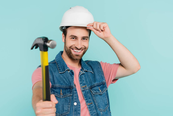 Positive man in denim vest and hardhat holding blurred hammer isolated on blue  - Фото, изображение
