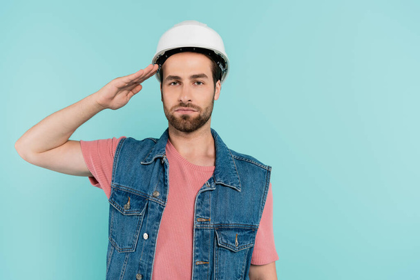 Young man in hardhat saluting and looking at camera isolated on blue  - Foto, Imagen
