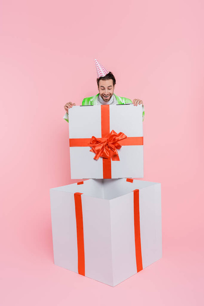 Excited man in party cap looking at huge gift box on pink background  - Zdjęcie, obraz