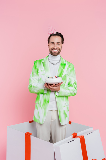 Positive man in party cap holding birthday cake while standing in gift box isolated on pink  - Fotografie, Obrázek