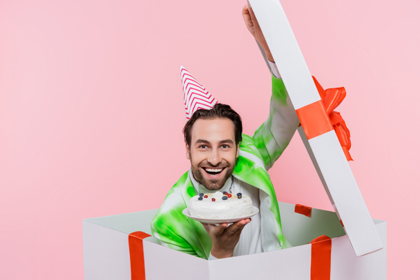 Веселий чоловік у вечірній кепці тримає торт на день народження, сидячи в подарунковій коробці ізольовано на рожевому
  - Фото, зображення