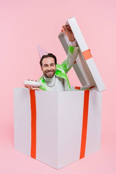 Happy man in party cap holding birthday cake while sitting in gift on pink background  - Foto, Bild