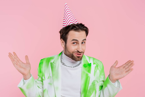 Young man in jacket and party cap gesturing and looking at camera isolated on pink  - Φωτογραφία, εικόνα