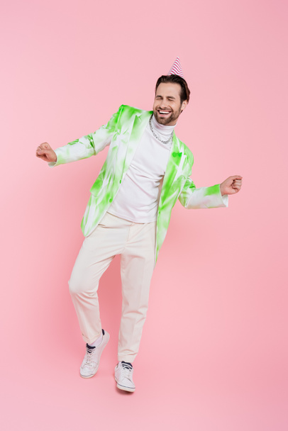 Cheerful man in party cap and jacket dancing on pink background  - Valokuva, kuva