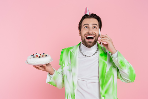 Uomo eccitato in berretto partito tenendo torta di compleanno e parlando su smartphone isolato su rosa  - Foto, immagini